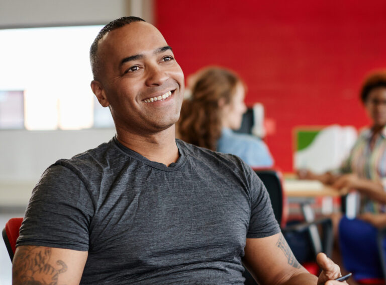 Casual portrait of an african american business man using technology in a bright and sunny startup with the team in the background